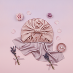 A pure silk sleeping bonnet, centered on a soft gradient background of blush pink, pale mauve, and soft lavender.