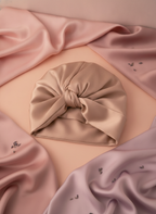 Close-up flat-lay of a pure silk sleeping bonnet on a warm surface with blush pink and pale mauve fabrics and lavender petals