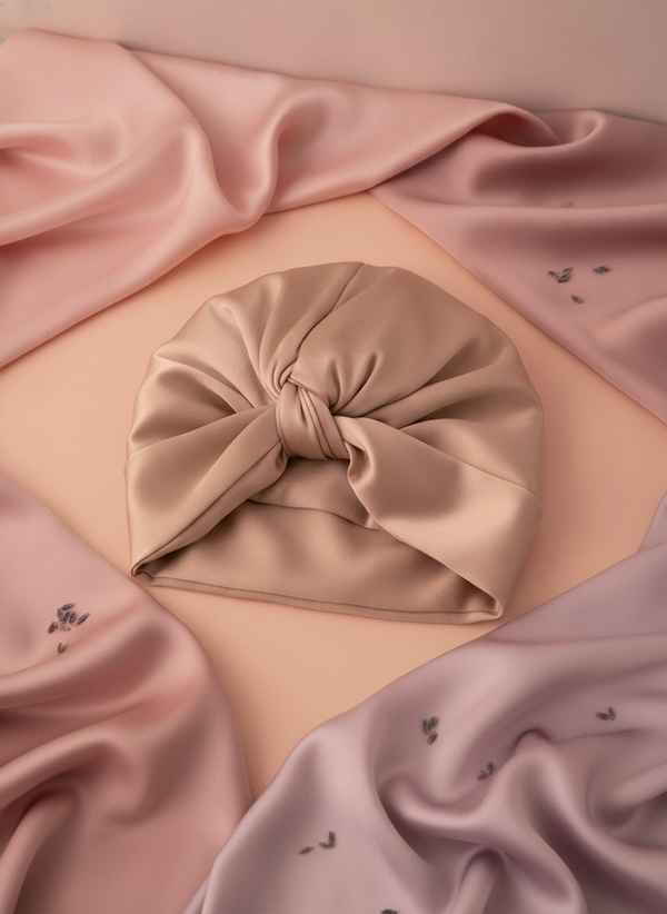Close-up flat-lay of a pure silk sleeping bonnet on a warm surface with blush pink and pale mauve fabrics and lavender petals